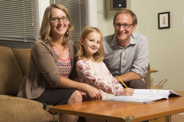Posed family photo, mother and father sit at table with daughter in front of them filling out a workbook. 
