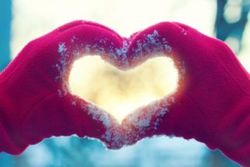 Red-gloved hands forming a heart shape in fresh snow, symbolizing holiday giving and community support.