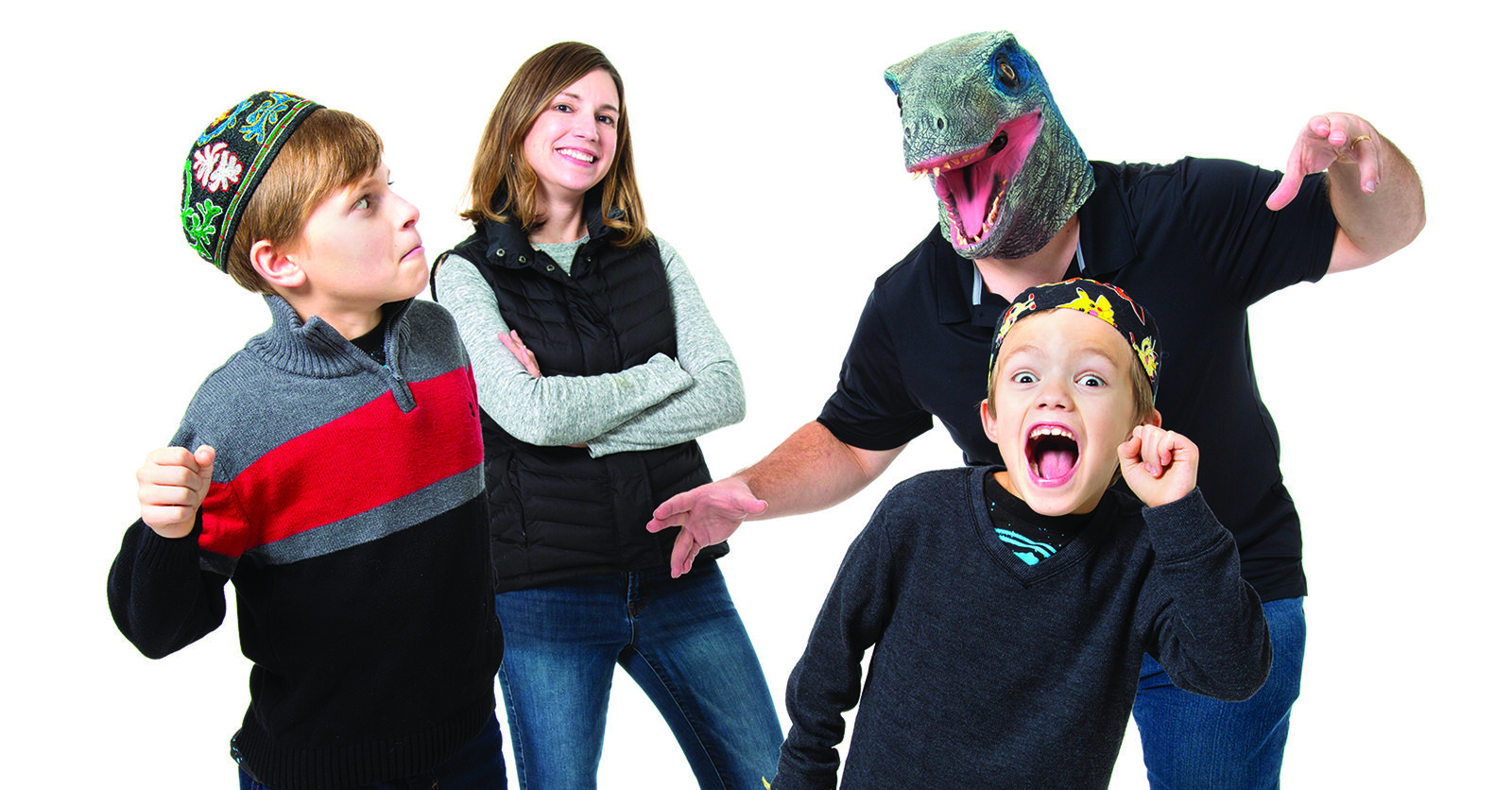 A playful family Hanukkah moment: two parents and their two sons smile and pose while the dad wears a goofy dinosaur mask, everyone laughing and having fun together.