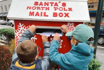 Two children place handwritten letters into a red “Santa’s North Pole Mail Box,” surrounded by holiday decorations.
