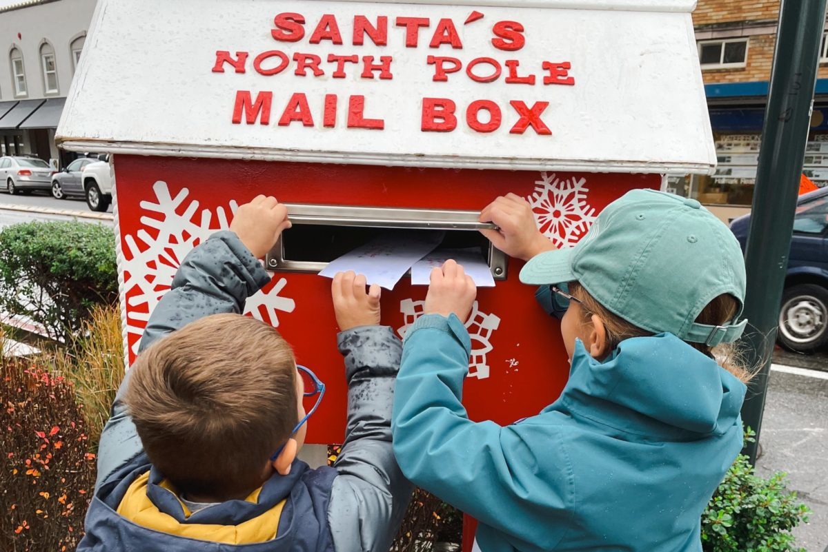 Two children place handwritten letters into a red “Santa’s North Pole Mail Box,” surrounded by holiday decorations.