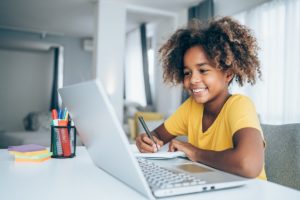 Child smiling and holding a pen while looking at a laptop screen
