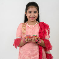 Young girl in traditional Indian dress proudly holding her colorful Diwali paper craft, surrounded by bright festive decorations.