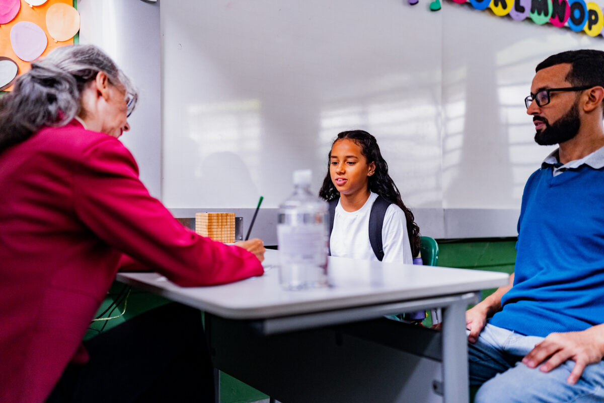 Teacher, parent and student sitting together at a classroom table reviewing student work during a parent-teacher conference.