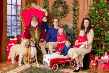 A family with four children and two dogs pose for a holiday photo with Santa, all smiling together in front of festive decorations.