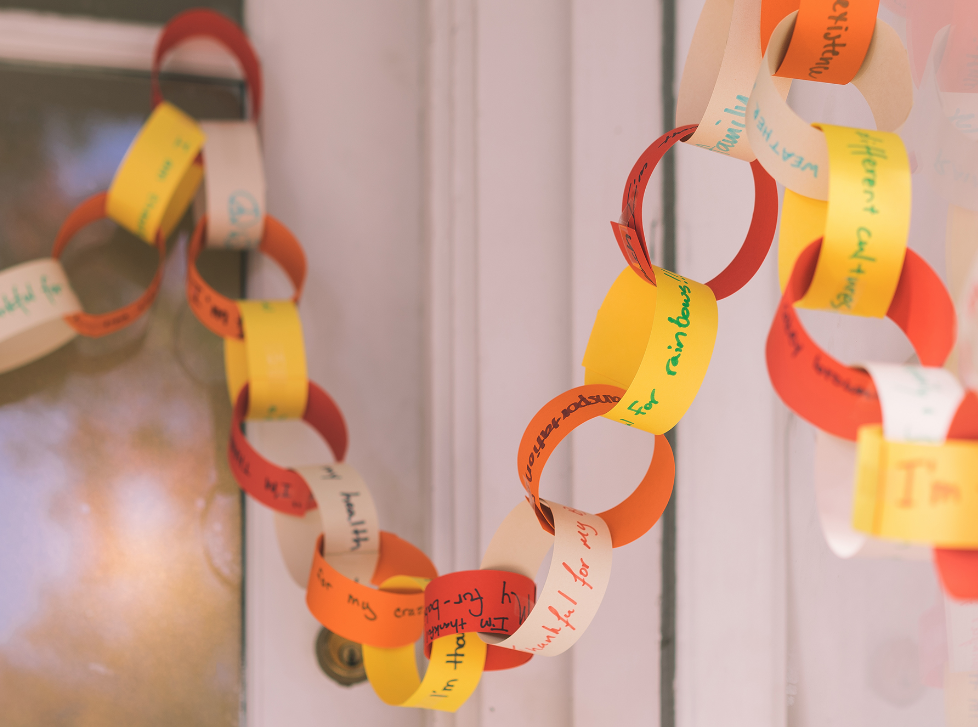 Close-up of a colorful paper “thankful chain” with handwritten gratitude messages linked together and draped across a living room shelf.