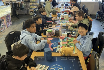 A group of children sit around a table at Tabletop Village in Seattle, playing Pokémon card games during a family game night.