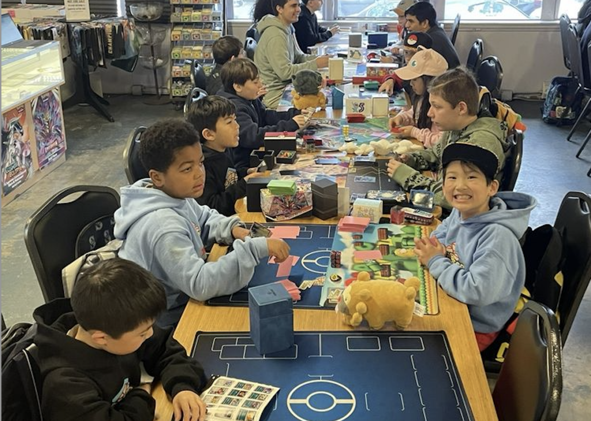 A group of children sit around a table at Tabletop Village in Seattle, playing Pokémon card games during a family game night.
