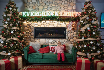 Girl sitting on a couch in front of a decorated fireplace with stockings and Christmas trees at Evergreen Christmas Lights in Bothell.