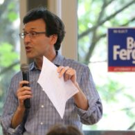 Bob Ferguson holds a mic and piece of paper mid-sentence, in front of a window with a Bob Ferguson election sign on it.