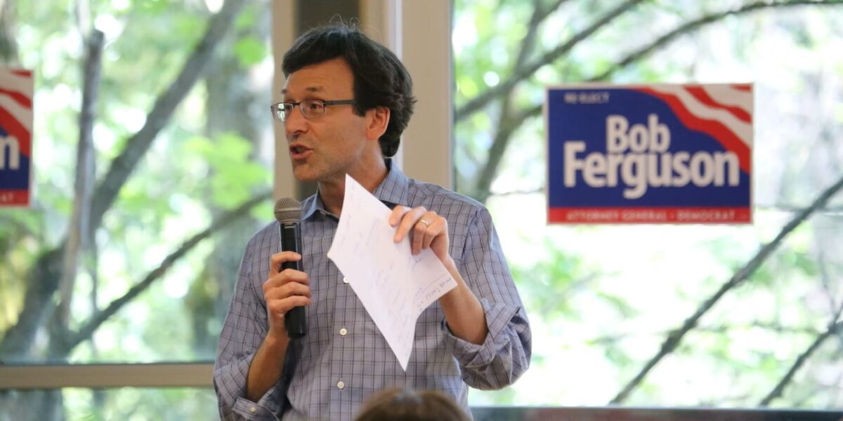 Bob Ferguson holds a mic and piece of paper mid-sentence, in front of a window with a Bob Ferguson election sign on it.