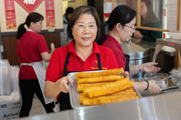 Woman behind counter at restaurant holds out plate of fried dim sum items, smiling at camera.