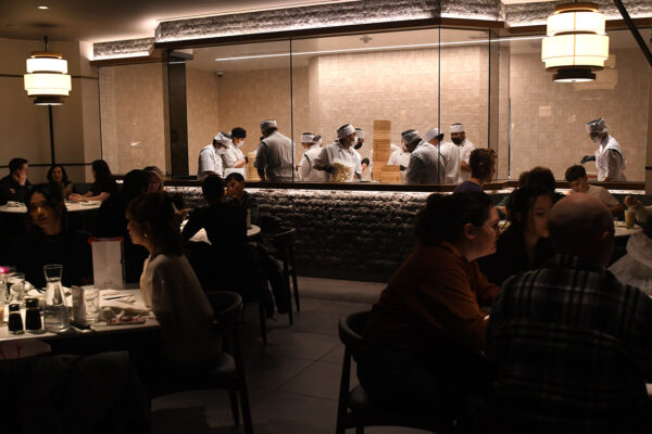 Interior of a dimly lit restauraunt packed with patrons. In the distance behind a glass window, you can see 10+ chefs bent over making dim sum.