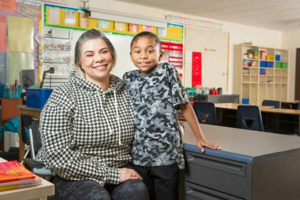 Woman sits in a kid's classroom with an elementary school kid posed next to her.