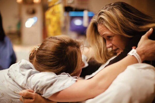 Woman in labor leans forward to hold onto another woman, her midwife. They are looking each other in the eye and holding tightly.