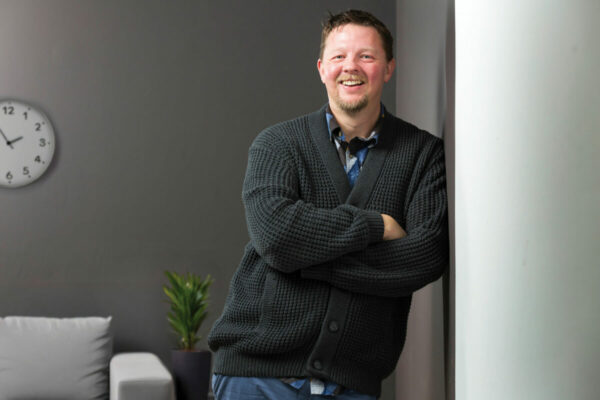 Man leans agains the wall of a sparse office, smiling at camera.