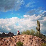 Boy with a red cap sits with his knees tucked in atop a desert hill, next to a cactus and under a bright blue sky.