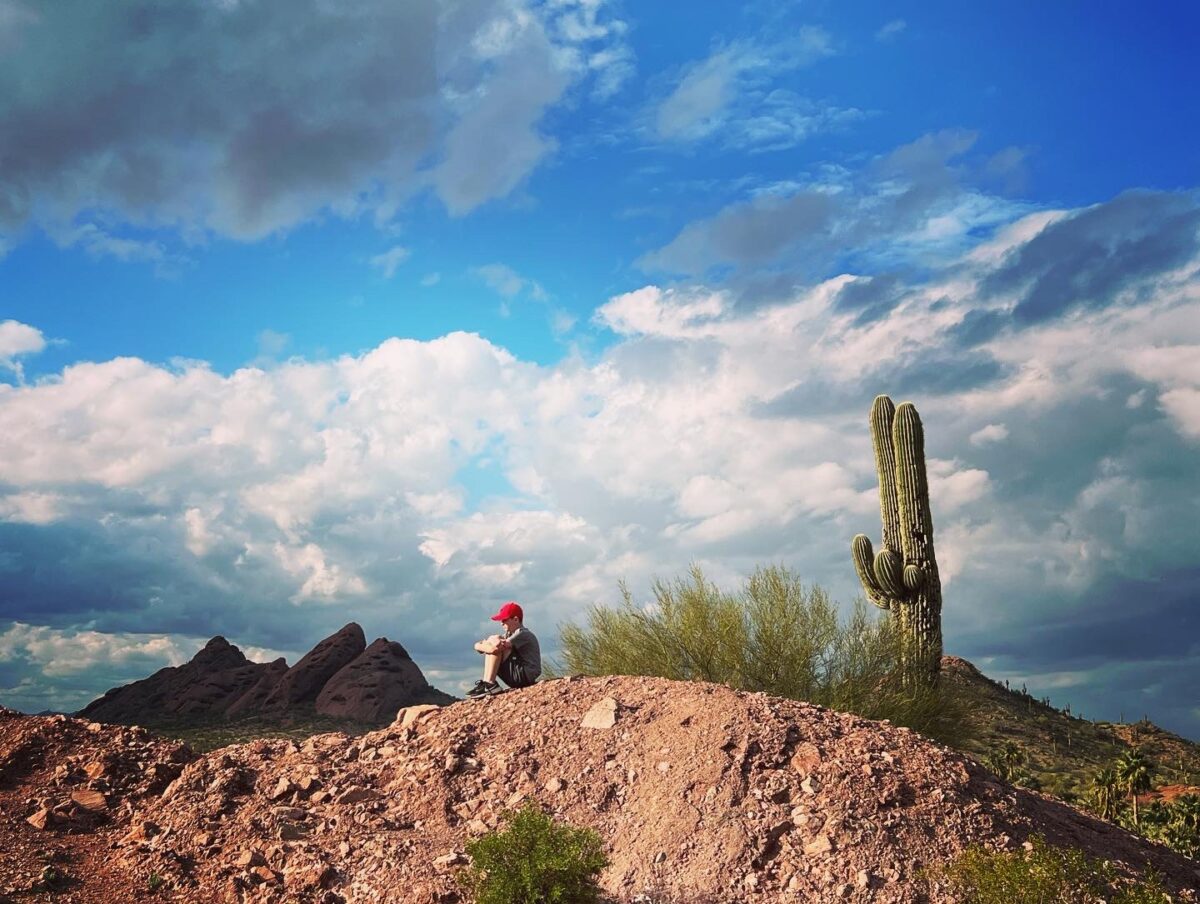Boy with a red cap sits with his knees tucked in atop a desert hill, next to a cactus and under a bright blue sky.