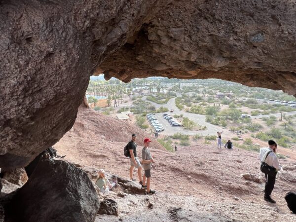 Hikers underneath a desert rock formation, with views of city in the distance.
