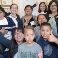 Group of kids and adults all huddle together facing the camera with thumbs up signs.