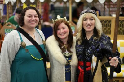 Three women smile at the camera, standing close together in medieval garb and crowns, holding scepters.