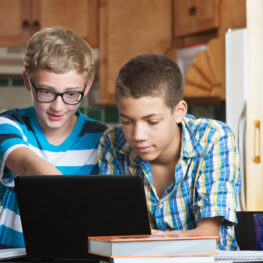 Two male teen friends in kitchen with books and laptop
