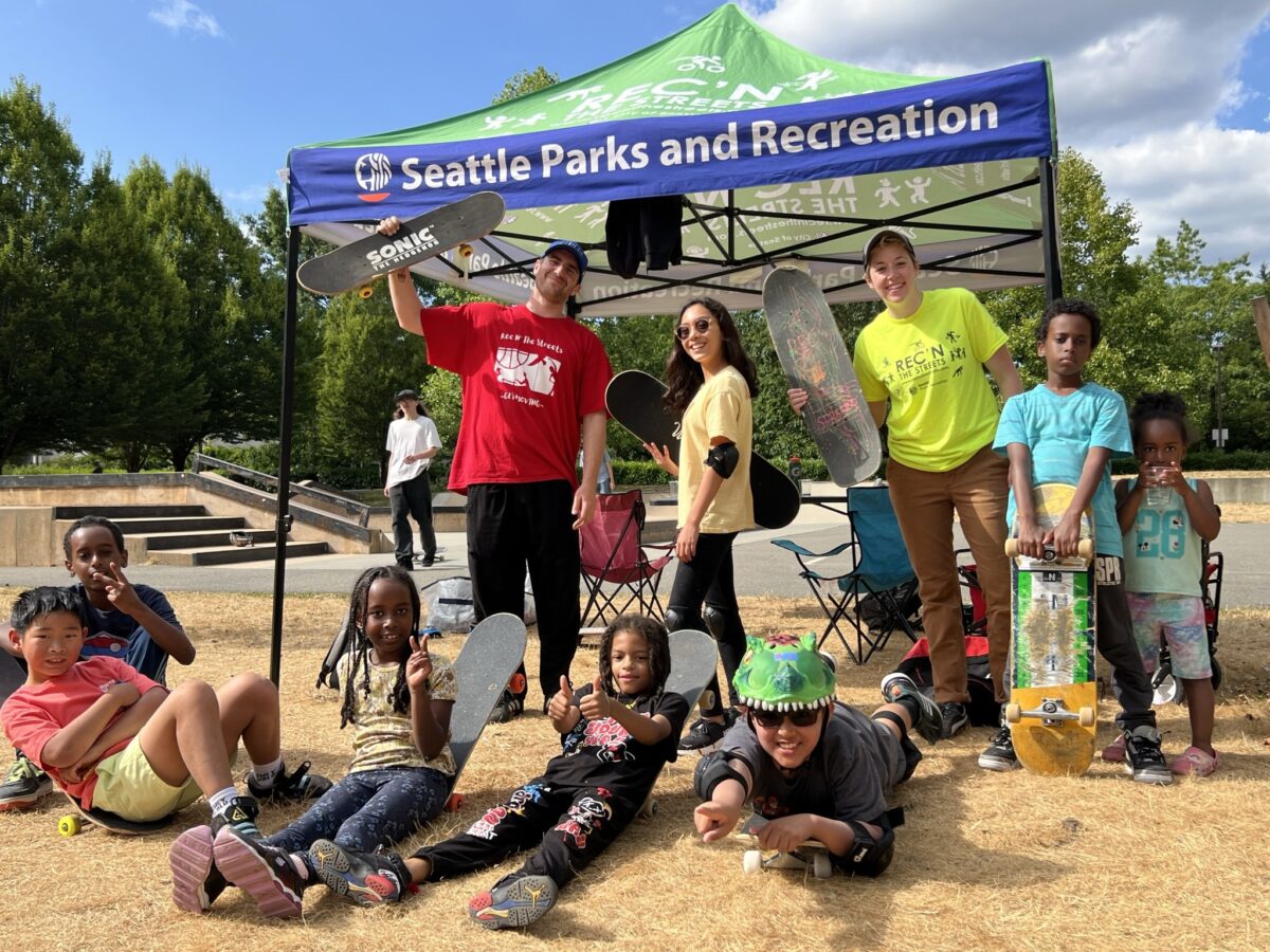 Kids and adults sit in front of tent that says Seattle Parks and Rec, dressed in skateboarding gear and holding boards.