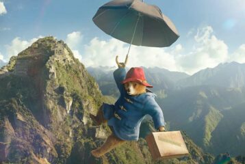 Paddington Bear flies through air with suitcase and umbrella, mountainscape in background