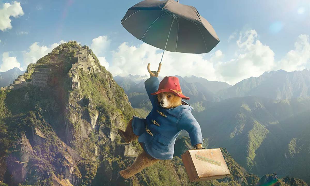 Paddington Bear flies through air with suitcase and umbrella, mountainscape in background