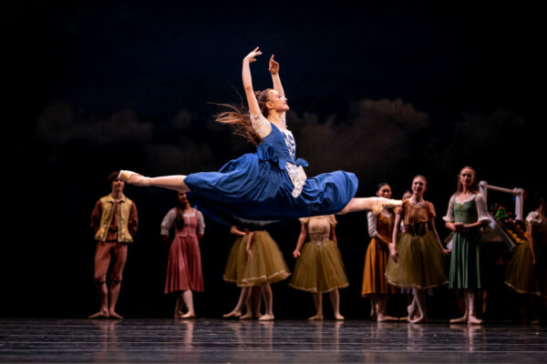 10 dancers in background stand watching single dancer in blue dress in foreground, doing the splits in the air