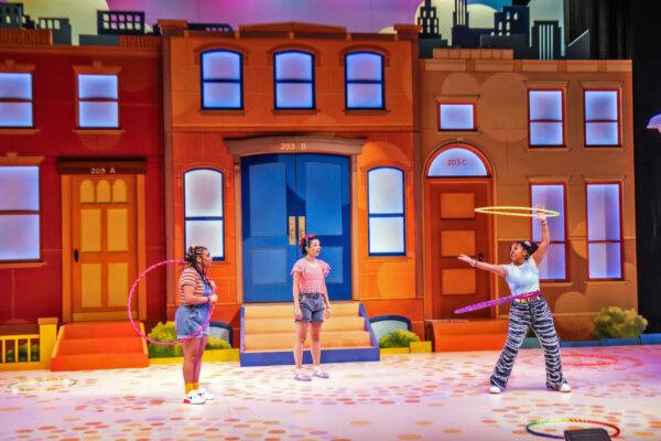 Three actors twirl hula hoops on a stage set to look like the block of a street. 