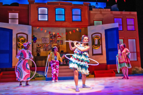 Actor hula hoops while playing the flute on stage