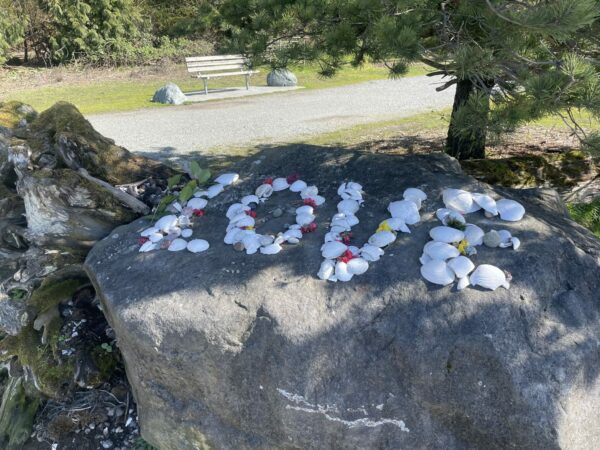 The word 'LOVE' spelled out with driftwood, rocks, kelp, flowers, crabshells, pinecones. Sitting on a large boulder in the park