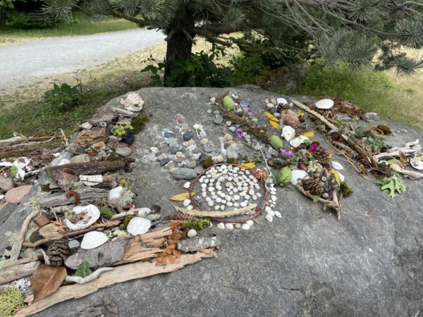 The word 'LOVE' spelled out with driftwood, rocks, kelp, flowers, crabshells, pinecones. Sitting on a large boulder in the park