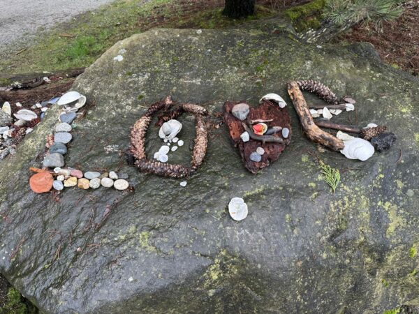 The word 'LOVE' spelled out with driftwood, rocks, kelp, flowers, crabshells, pinecones. Sitting on a large boulder in the park