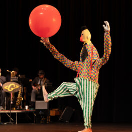 Costumed and masked performer wearing clownish costume hold big ball with foot up in kicking motion.