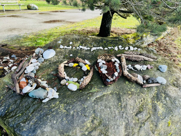 The word 'LOVE' spelled out with driftwood, rocks, kelp, flowers, crabshells, pinecones. Sitting on a large boulder in the park
