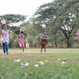 Happy group of diverse cute little children hunting Easter eggs, wearing bunny ears. kids holding basket, running to collect eggs on grass while playing outdoors at park
