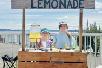 Etta, 10, and Hugo, 8, run a homemade lemonade stand, continuing a family tradition with the same wooden stand their dad used as a child.