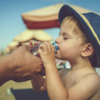 Adult helping child stay hydrated by offering a drink of water during hot weather.