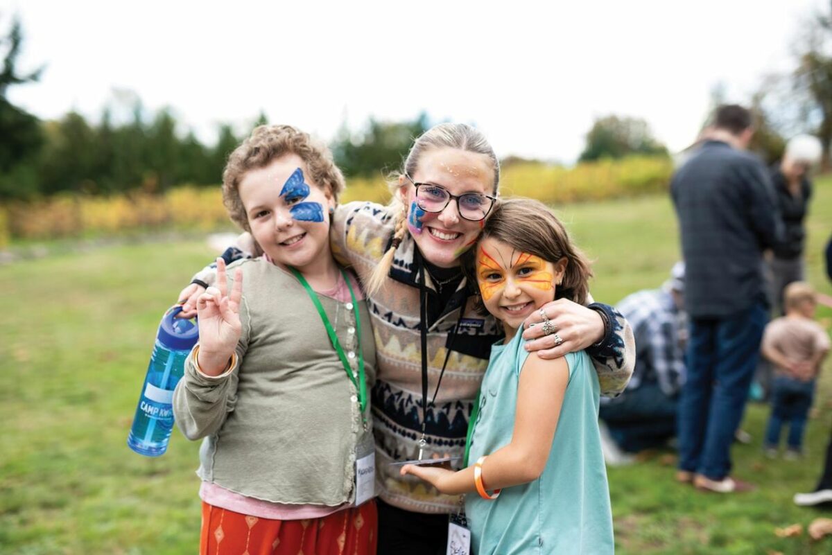 Two children and an adult share a warm hug at a Light Collective gathering, smiling with colorful face paint and surrounded by a joyful, welcoming atmosphere.