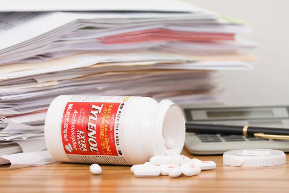 A tipped over and spilled bottle of Extra Strength Tylenol Acetaminophen Caplets in front of a pile of paperwork, a calculator, a pen, and the bottle lid. The Tylenol bottle contains 100 caplets, 500 mg each