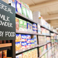 Close-up of a grocery store aisle with shelves of cereal and baby food, and a sign reading “Cereal, Milk Powder, Baby Food,” representing WIC-eligible items.