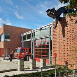 Exterior of Seattle’s Fire Station 9 with bright red bay doors and a large black-cat sculpture crouched on the rooftop, overlooking the sidewalk as a firefighter walks nearby.