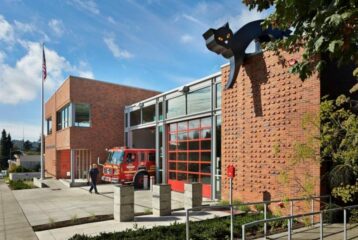 Exterior of Seattle’s Fire Station 9 with bright red bay doors and a large black-cat sculpture crouched on the rooftop, overlooking the sidewalk as a firefighter walks nearby.